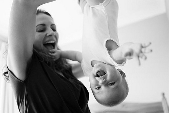 A mother playfully dangles her laughing baby by the foot at home.