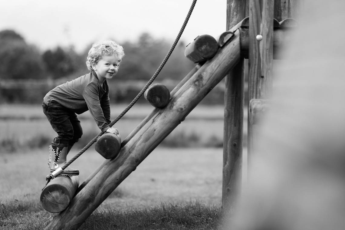 A black-and-white shot of a little boy at the park climbing a wooden climbing frame. Photo by Helen Bartlett.