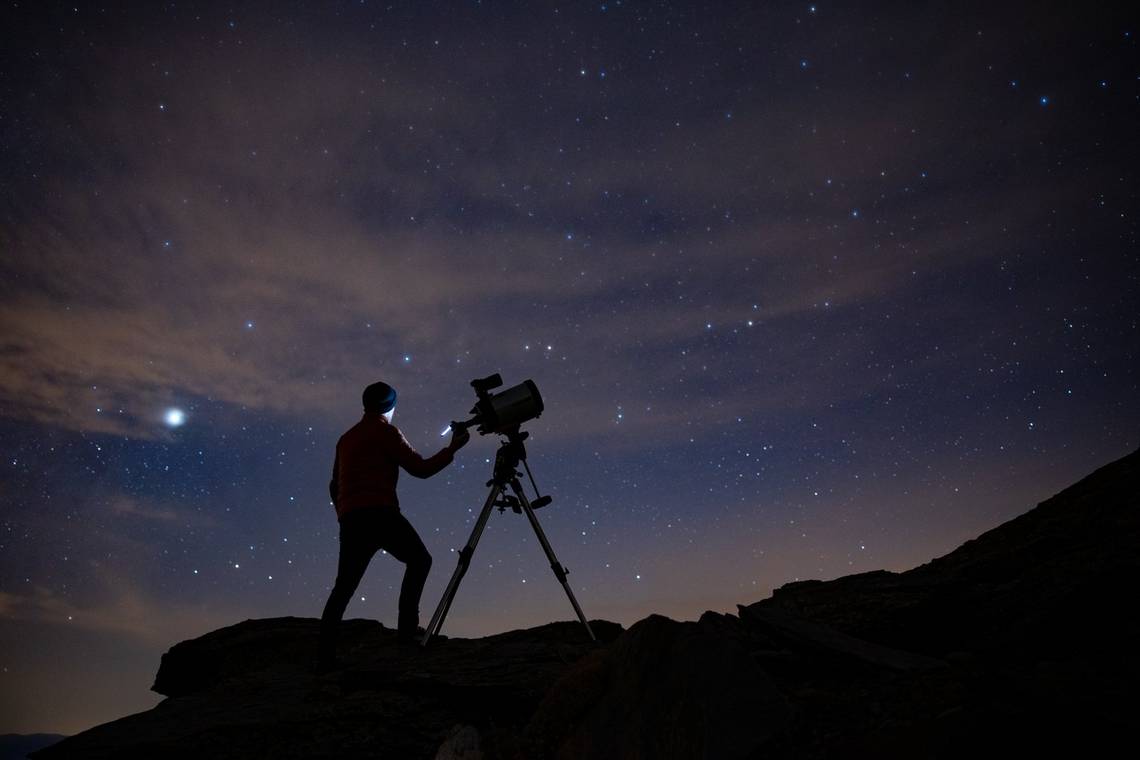 Silhouetted against the night sky, an astrophotographer stands on steep terrain with a Canon ֽ_격-a attached to a telescope.