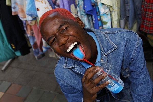 A young man drinking through a straw and sticking his blue tongue out.