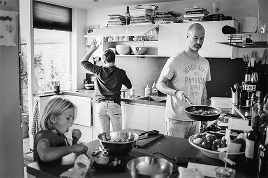 A man wearing t-shirt and jeans, frying pan in hand, prepares a meal in a bright kitchen. A teenage boy reaches up to a cupboard in the background, while an 8-year-old girl eats something in the foreground.