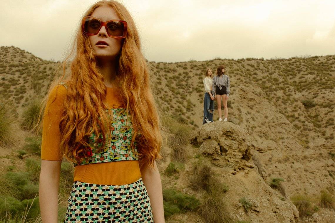 A female model stands outside in the foreground, with two other female models standing on a hill in the background.