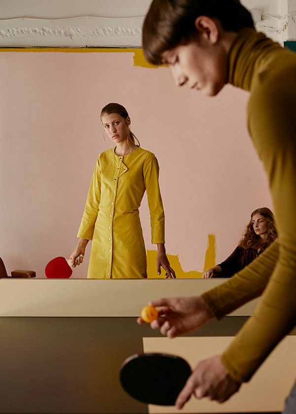 A man holds a ping pong bat and ball, looking intently down as if to start playing, and in the background a woman stands in a yellow dress, bat in hand.