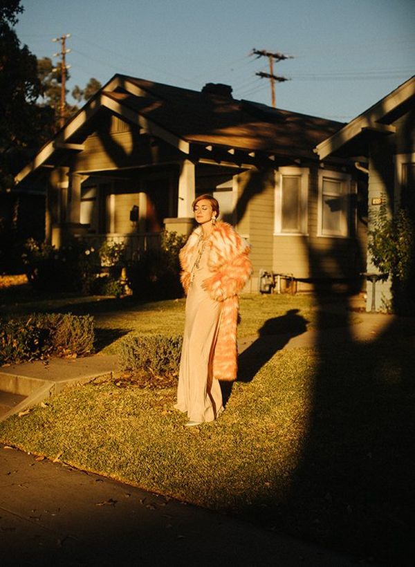 A woman stands in the front garden of a single-storey clapboard house, casting a long shadow as she faces a sunset.