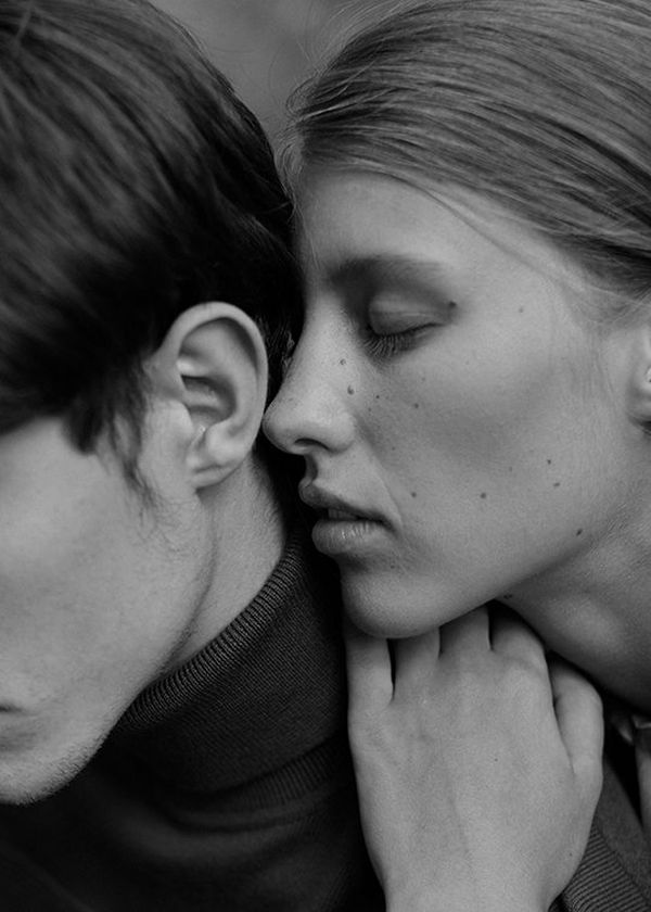 A black and white shot is composed with a young man mostly out of frame, his jaw and ear in shot, as a woman leans close to whisper to him.