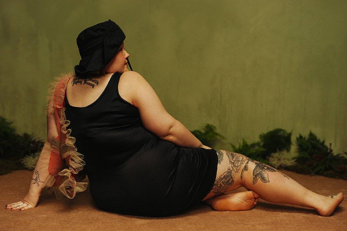  Plus-size, tattooed model Ami is photographed from behind, sitting with her legs to one side in a pose reminiscent of Old Master paintings of reclining models.