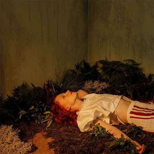 A redheaded woman wearing a silky off-white blouse and red and white striped trousers lies on her back on a bed of greenery with the corner of the set visible behind.