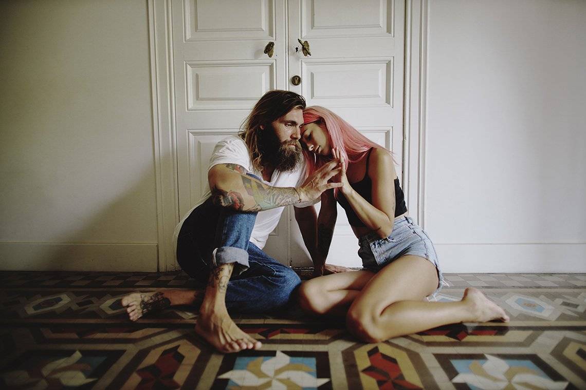 A long-haired, bearded man and a pink-haired woman sit on a tiled floor, their fingers intertwined.		