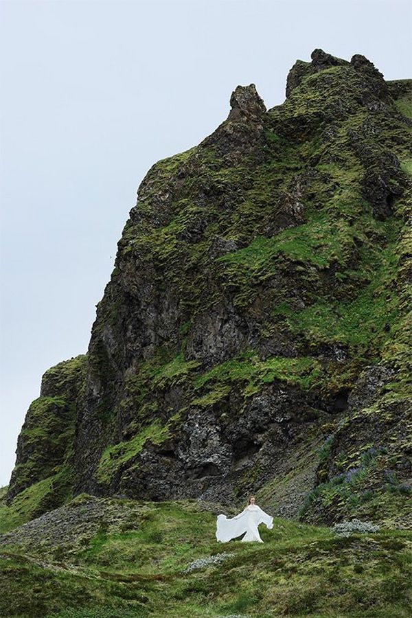 A woman in a flowing white wedding dress stands at the base of a steep, grassy rock face.