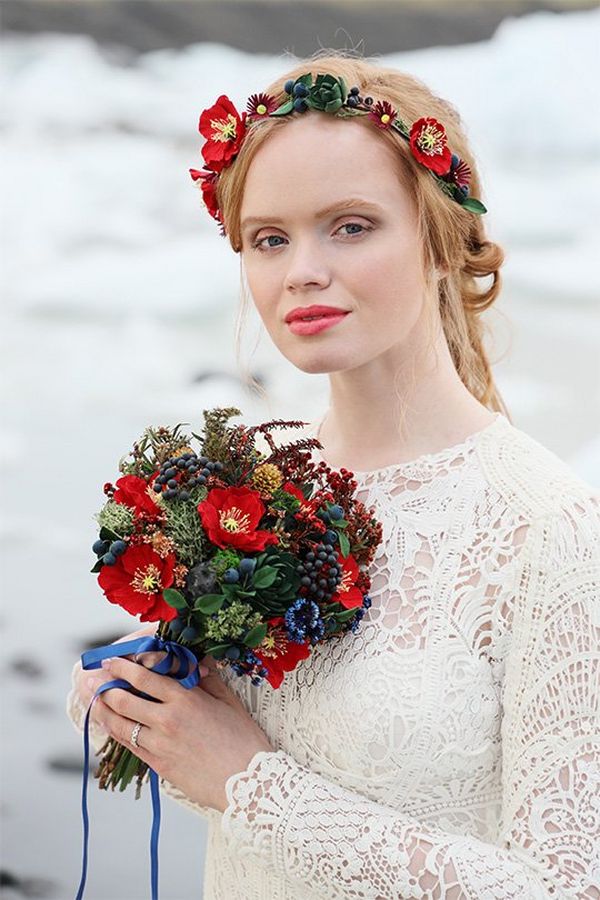 A strawberry blonde woman in a white lace wedding dress and flower headband holds a small bouquet of mixed red and blue flowers against an out-of-focus background of ice.