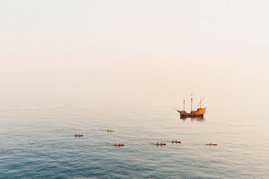 Six kayaks float in the water in front of a three-masted sailing ship with its sails furled; the sea vanishes in the glare, with no horizon visible.