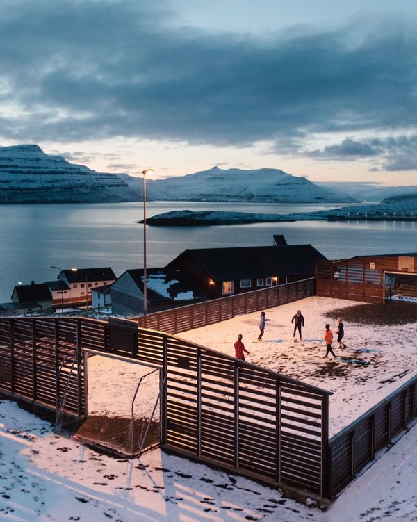 Five youngsters play football in a snow-covered ground inside a wooden fence; the waters of a chilly fjord are visible in the background.