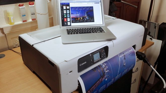 A laptop showing a 2024 calendar created on Canon PosterArtist sits atop a Canon large format printer. A print of the design is emerging from the printer. 