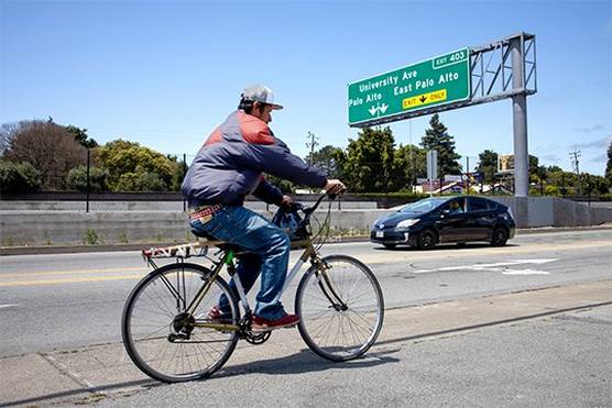 A cyclist and a car travel under a highway sign indicating the exit for University Avenue, Palo Alto and East Palo Alto. Photo by Laura Morton.