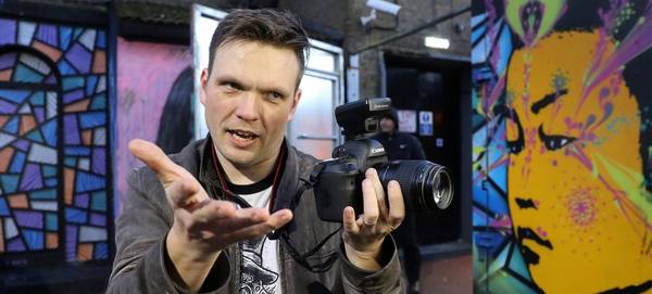 Photographer James Musselwhite holds a camera as he gestures to someone we can't see, in front of some street art.
