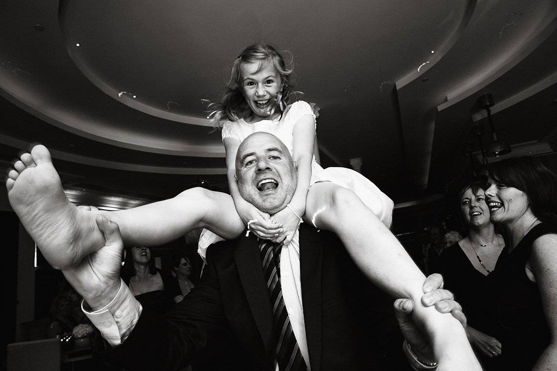 A man dances with his daughter on his shoulders, both laughing and looking at the camera.