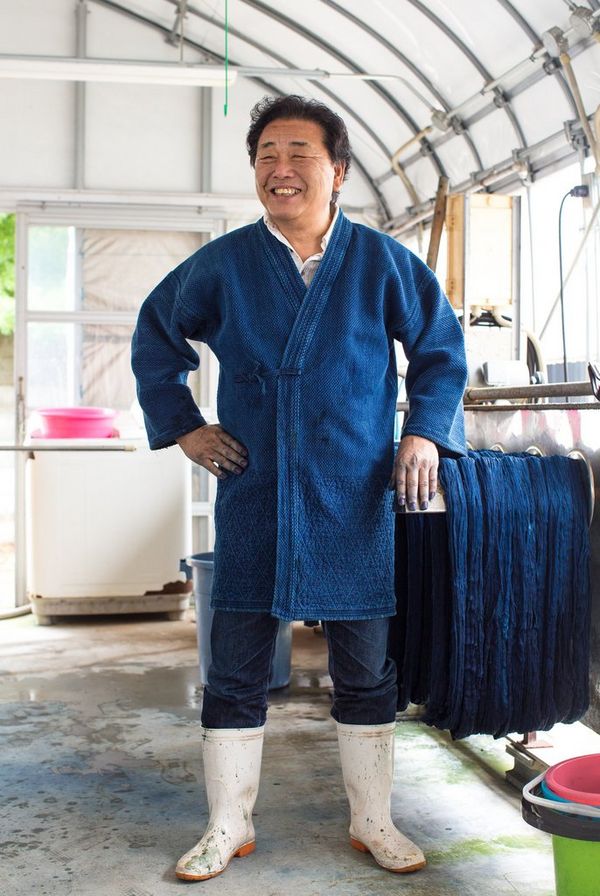 In a film still from Made in Japan, a smiling denim factory worker wears a blue denim robe, denim jeans and white boots.