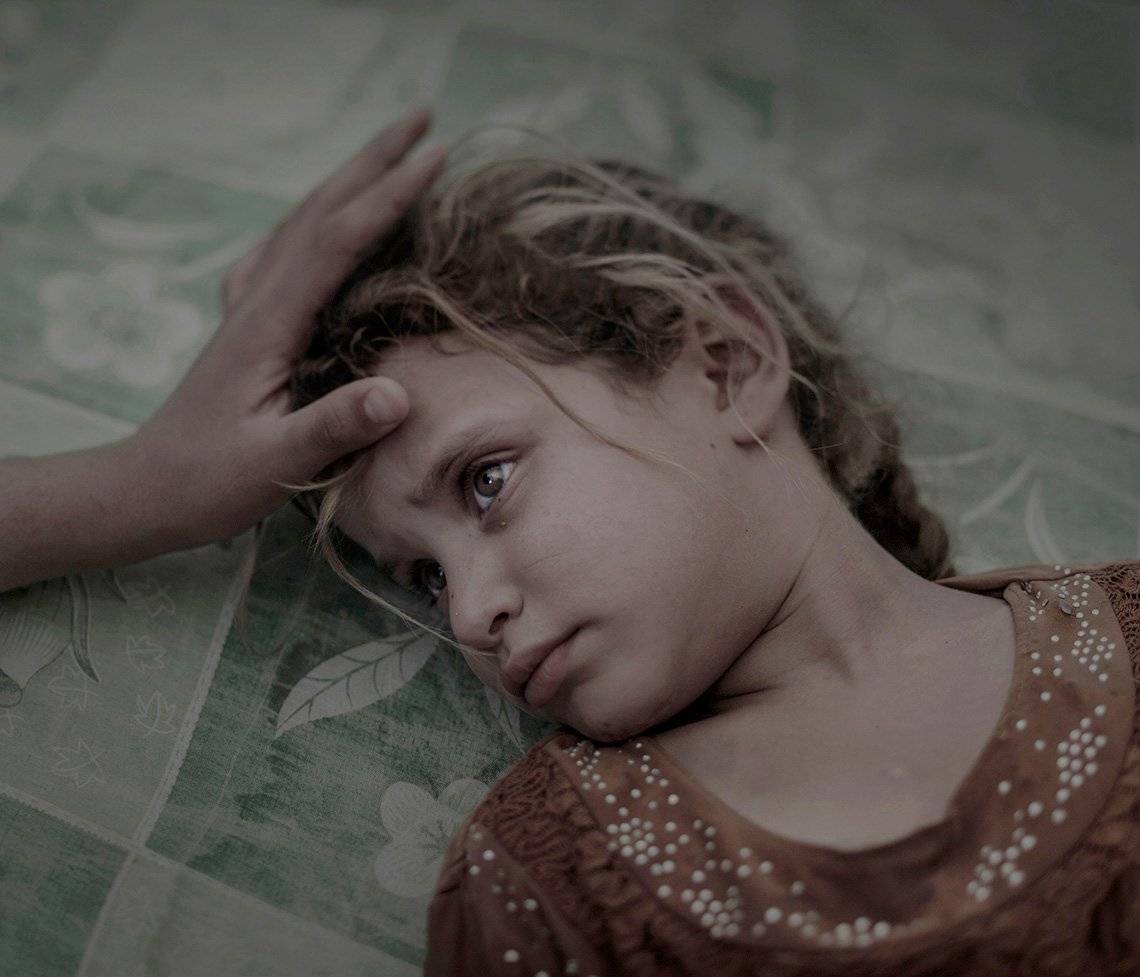 A blonde five-year-old girl lies on a green printed mattress, a hand stroking her hair