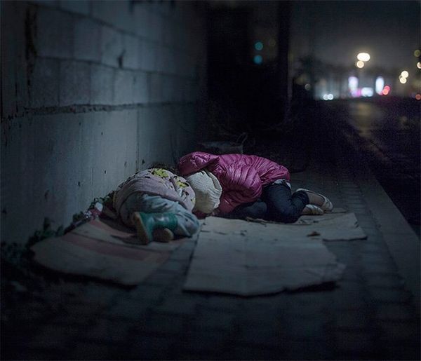 Two small children lie on flattened cardboard boxes by a wall in a dark, quiet street, wearing pink coats