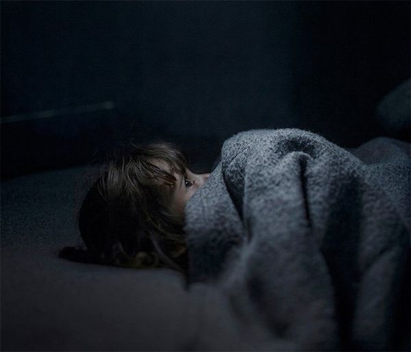 A small child lies on a grey bed under a grey wooden blanket that they have pulled up to cover most of their face. Their eyes are wide open, looking straight up at the ceiling