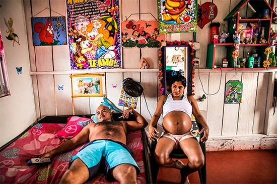 A heavily pregnant woman sits on a chair next to a man lying on a bed, her top rolled up to expose her stomach. They are in a brightly-lit room decorated with colourful posters and a shelf unit holding cosmetics bottles and small stuffed toys.