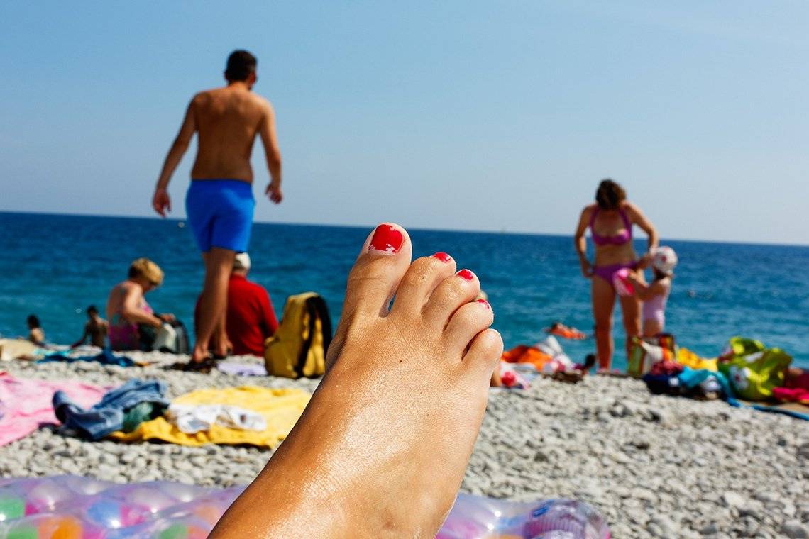 A beach scene. A woman's foot with bright red nail varnish is in the foreground, while families relaxing by the blue sea and skies are in the background.