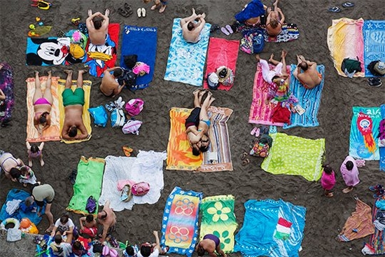 An overhead shot of people sunbathing on colourful towels at a black sand beach. Taken by Martin Parr on a Canon EF 70-300mm f/4.5-5.6L IS USM lens.