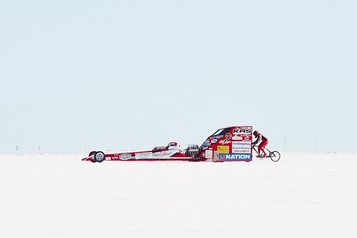 A racing car with raised sides at the back and a cyclist following along Utah's salt flats.