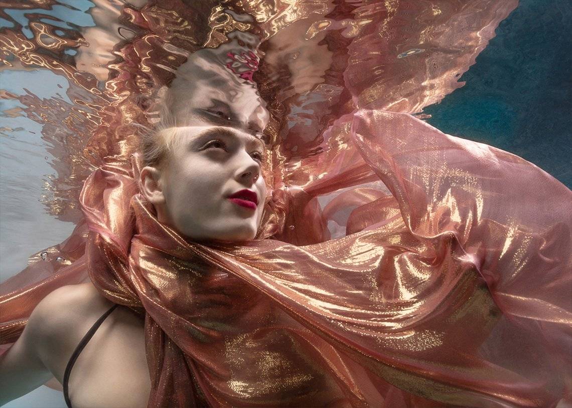 A woman submerged in water with a pink and gold scarf billowing around her.