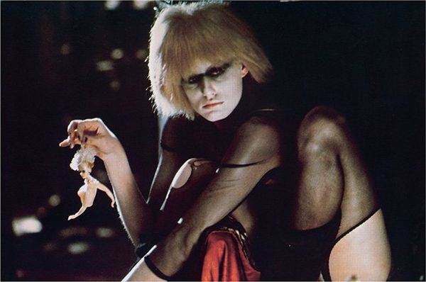 Daryl Hannah in costume as Pris from Blade Runner, wearing a sheer black leotard, heavy black eye makeup and holding a doll by the hair.