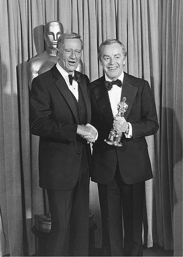 John Wayne and Michael Deeley shaking hands at the Oscars, Michael holding an award.