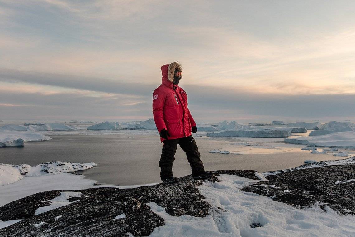 An Ariston installer wearing a red puffer jacket with a fur-effect hood, balaclava and eye protection stands on snow-covered rocks in front of a body of water strewn with icebergs. 