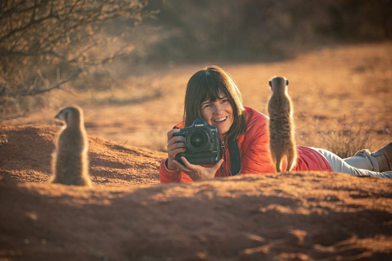 Wildlife photographer Marina Cano lying on the desert floor holding a Canon EOS-1D X Mark III in front of two baby meerkats.
