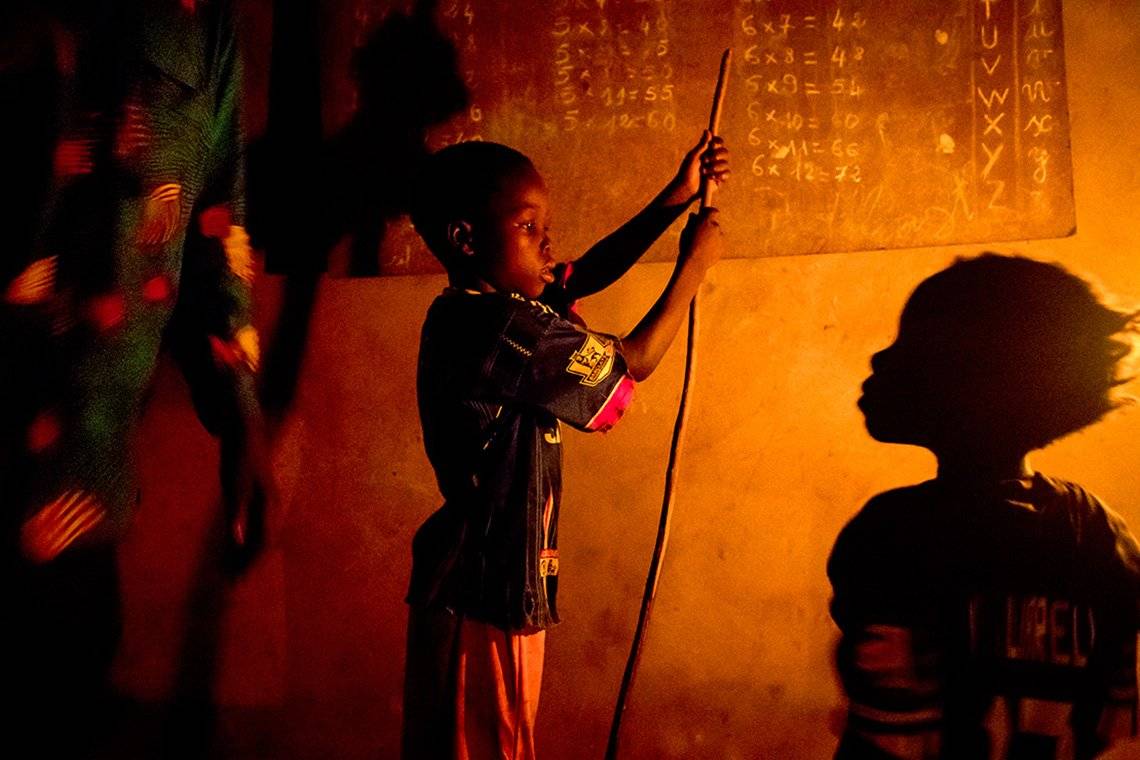 Two children stand in front of a blackboard at home in the village of Allankpon, Benin, practicing multiplication tables thanks to the orange light of a kerosene lamp.