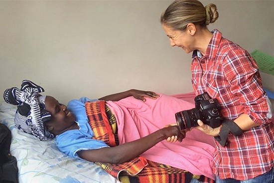 Photographer Georgina Goodwin, holding her Canon camera, greets a woman in a hospital bed in Kenya.