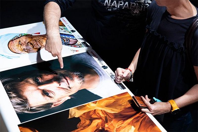 People look at three photos on a display - the left, of yellow flame-like flowing fabric, the middle, a close-up of a middle-aged man, the right of an older African woman in colourful printed clothes.