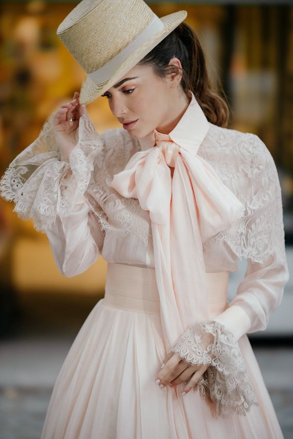A model in a frilly wedding dress and top hat, photographed against a brightly-lit shop front in evening light.
