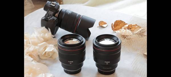 A Canon RF 85mm F1.2L USM lens and RF 85mm F1.2L USM DS lens side-by-side on a table, with a Canon EOS R behind.