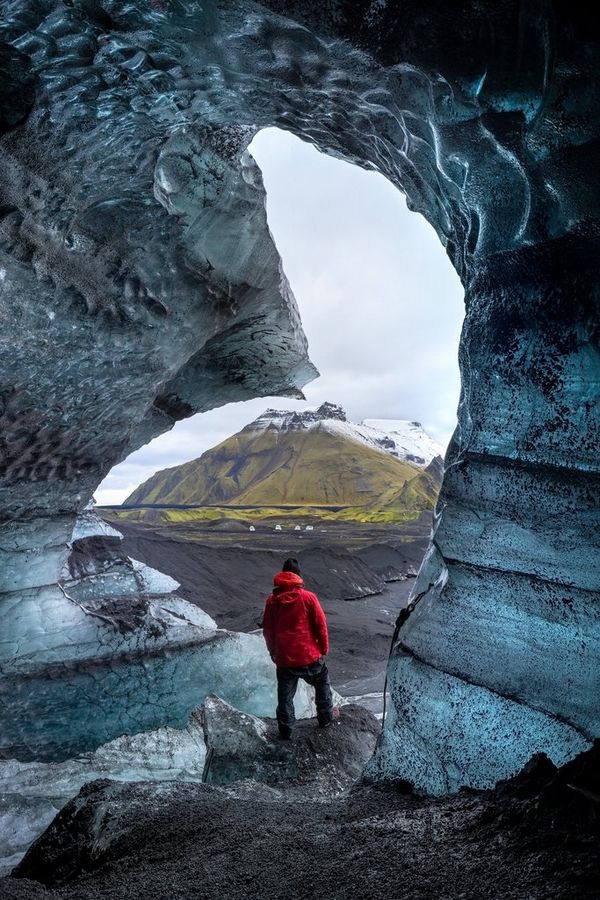 A man in a red padded coat stands at the mouth of an ice cave looking out at a bleak landscape with a snow-capped mountain in the distance.
