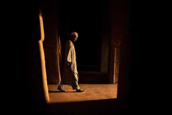 A man in flowing clothing and a cap walks barefoot through a dark room in Niger, the only light being from the doorway he's approaching.