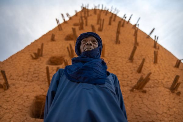 A man wearing an indigo robe and headscarf stands in front of a tower in Niger. Taken by Joel Santos.
