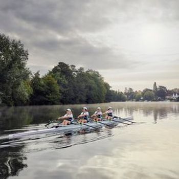 A crew of four women row a coxless four boat along a wide river at dawn. Photo by Matthew Joseph.