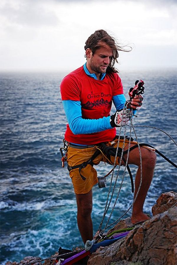 A man stands at a windswept cliff edge by the sea preparing ropes. 