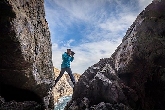 Action photographer Richard Walch shooting with a Canon ֽ_격-, one leg on each side of a steep coastal cliff.
