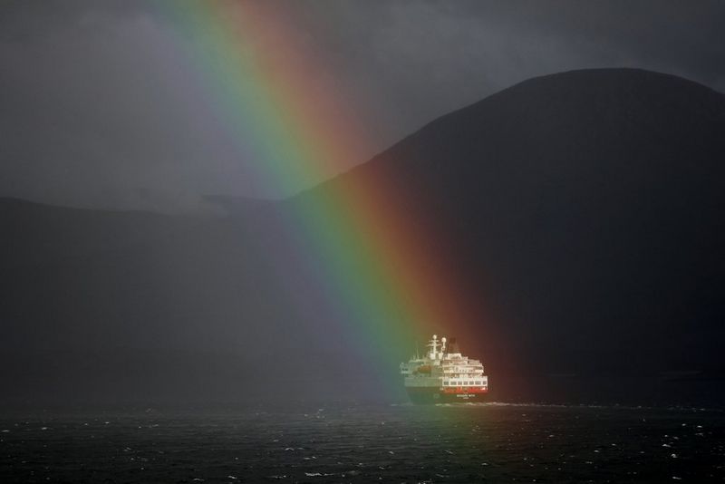 A striking rainbow illuminates a ship in an otherwise dimly-lit Norwegian fjord. Photo by Richard Walch.