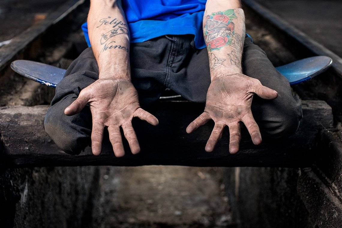 Double leg amputee skateboarder Felipe Nunes sits on a skateboard in a train garage while showing his hands, covered in dirt from being used to propel himself along during a skateboarding session. 