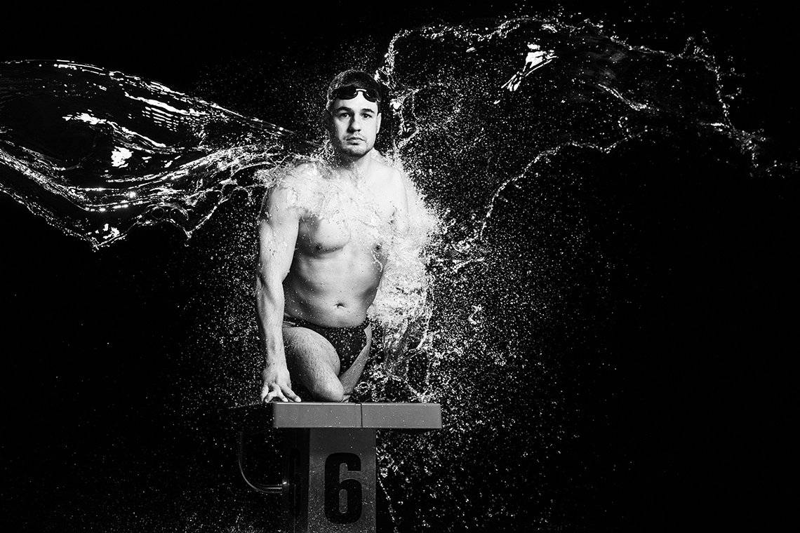 Disabled swimmer Darko Duric stands on a diving board without his prosthetic legs, wearing swimming briefs and goggles as if ready to dive. Two large splashes of water hit him from either side, forming wing-like shapes.