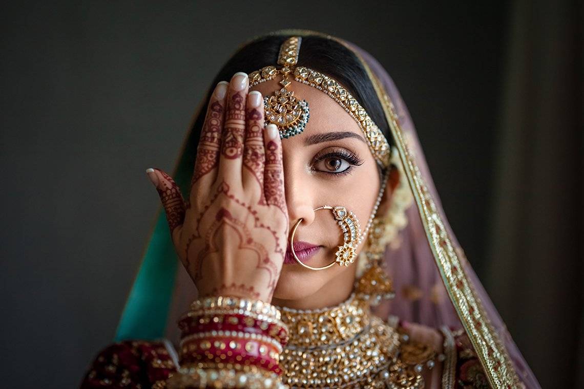 An Indian woman in silk wedding clothes and opulent gold jewellery holds a henna tattooed hand in front of one eye.