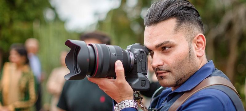 Wedding photographer Sanjay Jogia crouches low among some stones, holding his Canon camera to his eye.