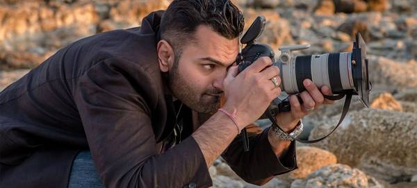 Wedding photographer Sanjay Jogia crouches down low on a beach, holding his Canon camera and long lens to his eye to photograph someone out of shot.